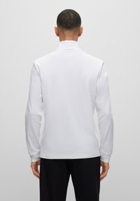 Camisa polo de manga larga en blanco, con un cuello alto y una textura suave, con dobladillo recto y detalles de diseño mínimos.