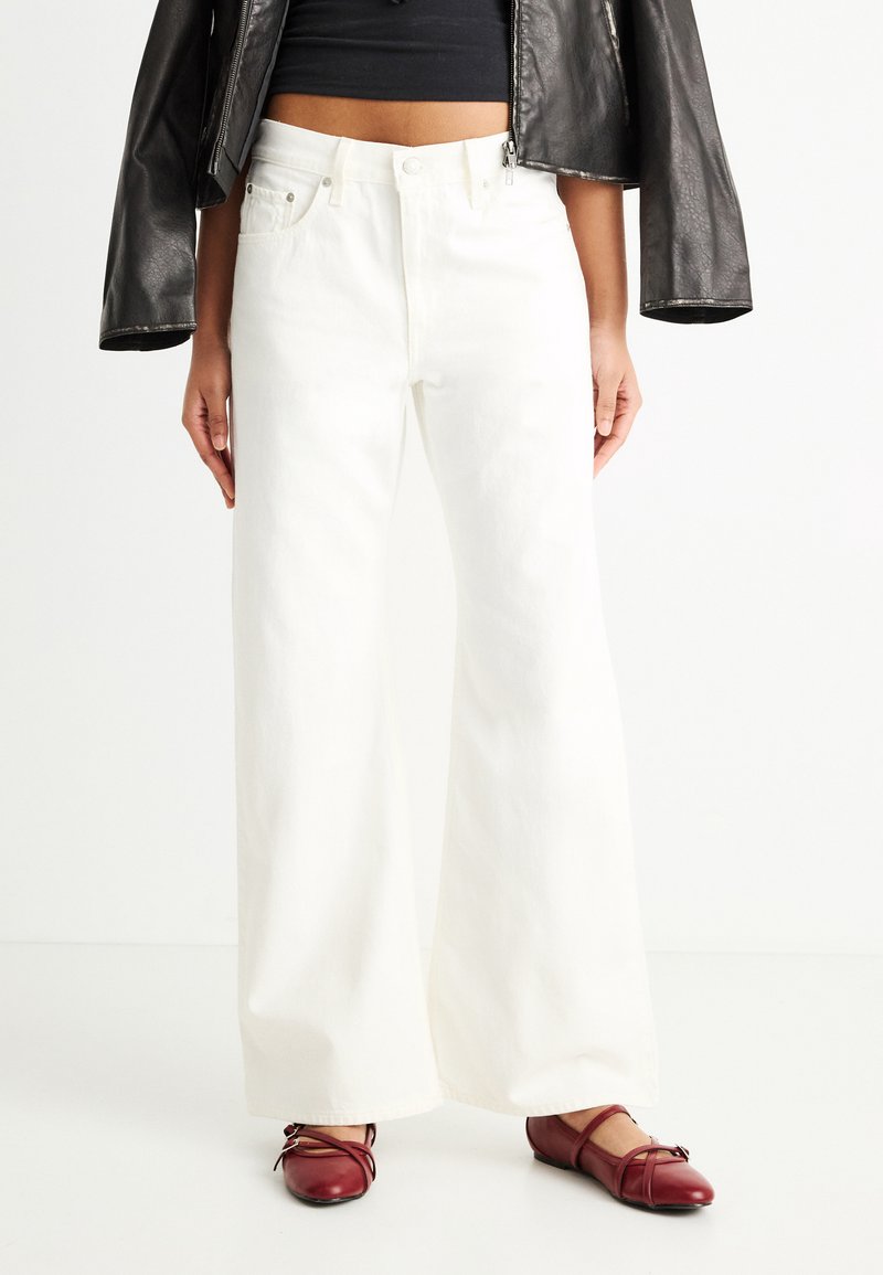 Άτομο που φοράει λευκό παντελόνι με ψηλή μέση και φαρδιά μπατζάκια, μαύρο cropped τοπ, μαύρο δερμάτινο σακάκι και κόκκινα επίπεδα παπούτσια με λουράκια στον αστράγαλο.