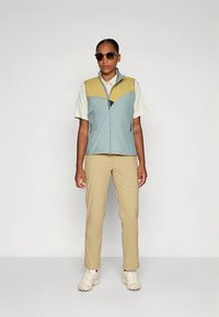 Leichte, mehrfarbige Weste mit einem blau-braunen Design, getragen über einem beigen Polohemd und kombiniert mit beigen Hosen und Sneakers.