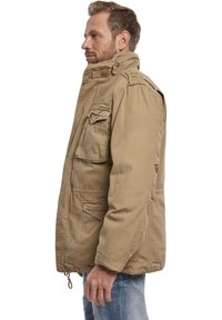 Jachetă utilitară khaki, cu multiple buzunare frontale, guler înalt și epauleți la umeri. Fabricată dintr-un material rezistent din bumbac combinat.