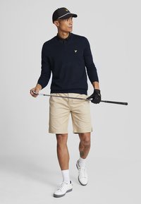 Navy v-neck sweater with yellow logo, beige shorts, black golf glove, white sneakers, and a black cap with white accents, holding a golf club.