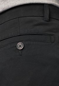 Pantaloni di cotone neri con una tasca posteriore dotata di un bottone argentato, cuciture ordinate e una texture liscia. La vita è piatta.
