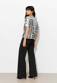 Haut en sequins argentés à manches courtes, associé à un pantalon large à carreaux noirs et blancs ainsi qu'à des chaussures à talons hauts. Fond blanc uni.