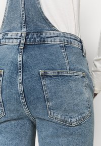 Siniset farkkuliivit, joissa on haalistunut pinta, säädettävät olkaimet ja kaksi takataskua tikatulla reunoilla. Kangas on pehmeää.