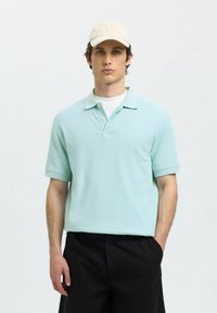 Ljusblå polotröja med korta ärmar, tre knappar och kontrasterande krage. Bärs med svarta shorts och en beige keps.