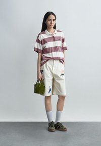 Femme portant un polo rayé mauve et blanc, des shorts crème brodés, des chaussettes blanches, des baskets olive, tenant un petit sac à main olive.