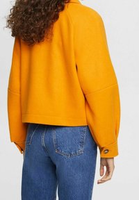 Orange wool-blend jacket with oversized sleeves, collar, and button details, paired with blue denim jeans, showing a relaxed fit and texture.