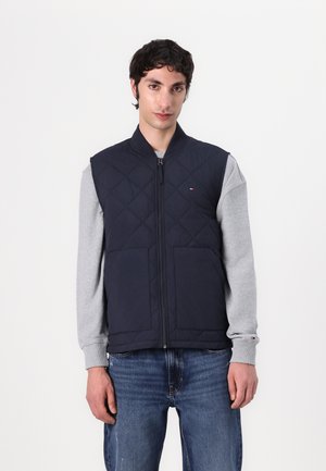 Gilet matelassé bleu marine avec fermeture éclair à l'avant, poches latérales et col montant, porté sur une chemise grise à manches longues.
