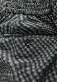 Tengerészkék és krémszínű, mintás szövet, egy zseben egyetlen fekete gombbal. Az anyag texturált, finom kockás mintával.