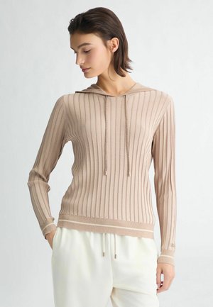 Sweat à capuche beige clair avec des côtes verticales, manches longues et cordons de serrage. Texture douce avec un léger brillant, associé à un jogging crème.