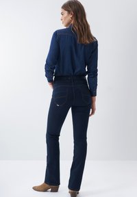 Mørkeblå denim flared jeans med baglommer og orange syninger, parret med en matchende blå langærmet skjorte. Beige stiletter.