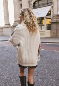 Chunky knit cardigan in cream with patterned accents on sleeves. Styled over a dark skirt and paired with knee-high black boots.