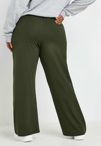 Olivgröna vida byxor i elastiskt tyg, med en slät textur och en enkel design. Bärs med en grå sweatshirt och vita skor.