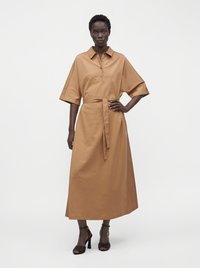 Femme portant une longue robe chemise couleur camel avec manches courtes, col boutonné, taille nouée, et des sandales noires à talons hauts.