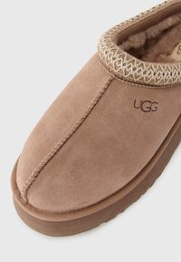 Brown suede slip-on slipper with soft wool lining and UGG logo on top, featuring decorative stitching around the ankle opening.