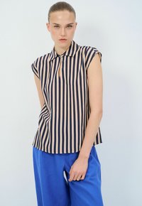 Kurzärmelige, gegliederte Bluse mit senkrechten dunkelblauen und hellrosa Streifen; Schlüssellochöffnung am Hals; kombiniert mit blauen Hosen.