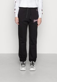 Černé tepláky z měkké látky se stahovací šňůrkou v pase a růžovým logem „Champion“ na levém stehně; doplněné masivními bílými teniskami.