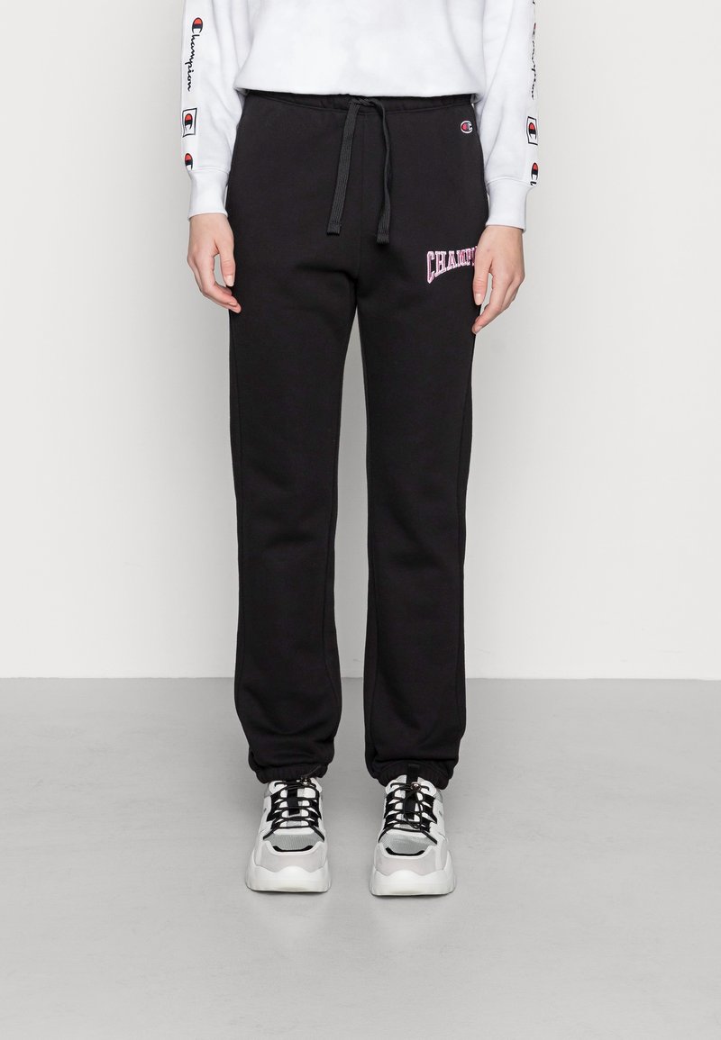 Svarta mjukisbrallor i mjukt tyg med dragsko i midjan och rosa "Champion"-logga på vänstra låret; kombinerade med grova vita sneakers.