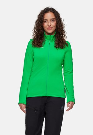 Kvinde med krøllet hår iført en grøn lynlåstrøje og sorte bukser, stående foran en ensfarvet lys baggrund.