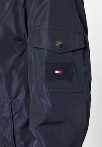Giacca in nylon blu navy con un design elegante, caratterizzata da una tasca a patta con un'etichetta nera che mostra "TOMMY HILFIGER" e colori dell'emblema in rosso, bianco e blu.
