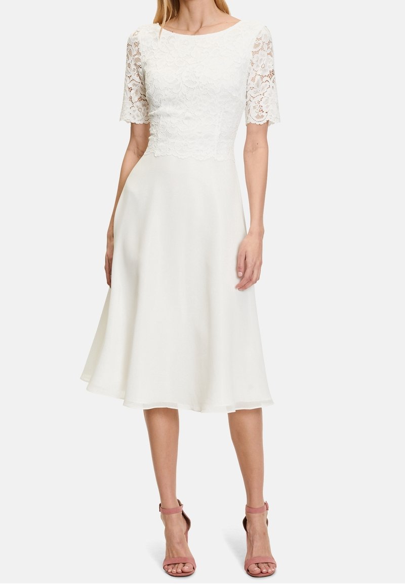 Femme portant une robe blanche mi-longue avec manches courtes en dentelle et corsage, associée à des sandales roses à talons hauts, debout sur fond blanc.