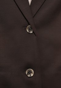 Brūns auduma žakete ar izgrieztu atloku un divām pamanāmām pogām ar smalku spīdumu. Texturēts materiāls ar gludu virsmu.