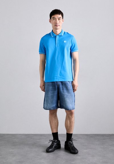 Cămașă polo albastră cu guler în dungi, asociată cu șorturi din denim. Modelul poartă adidași negri și șosete, stând pe un fundal neutru.