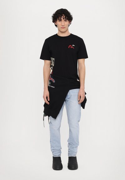 T-shirt noir avec broderie rouge et détail graphique sur le côté, associé à un jean clair et des bottines noires à talons. Sweatshirt noir drapé autour de la taille.