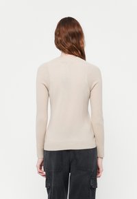 Beige ribbestrikket genser med lange ermer, rund hals og tettsittende midje, laget av mykt strikket stoff, sett fra bak.