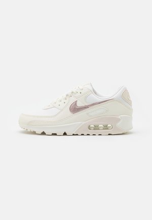 Niski, biały sneaker z beżową podeszwą i fakturowanym jasnoróżowym logo Nike, widoczną amortyzacją powietrzną i sznurowanym fasonem, widoczny z boku.