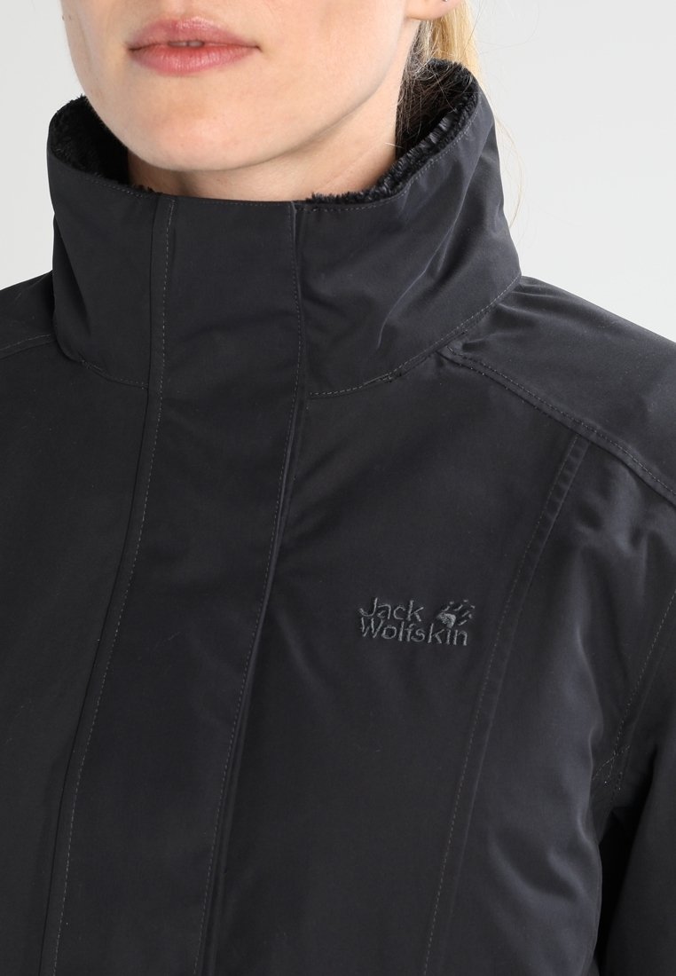 Femme portant une veste noire Jack Wolfskin à col montant, montrant le logo brodé sur la poitrine, avec les lèvres et une partie du cou visibles.
