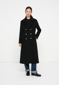 Manteau noir croisé avec de grands boutons argentés, poches latérales et col cranté, fabriqué en tissu structuré, avec une coupe mi-longue.