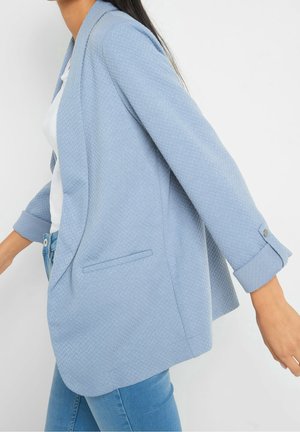 Světle modrý texturovaný blazer s uvolněným střihem, dlouhými rukávy a jednou přední kapsou. Rukávy mají knoflíkové detaily. Nošený přes bílý top.