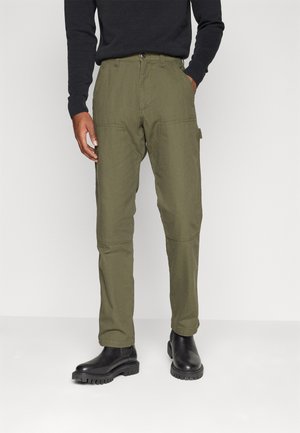 Pantalons cargo vert olive avec une coupe droite, en coton. Comprend des poches latérales et des genoux renforcés. Portés avec des bottines noires.