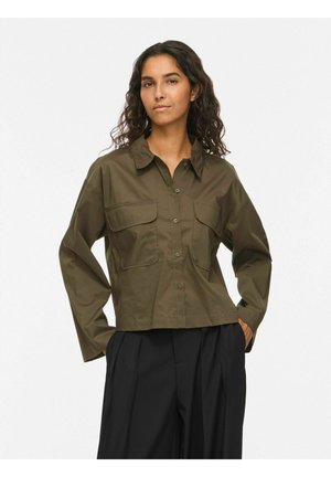 Femme aux cheveux bouclés foncés portant une chemise verte olive boutonnée avec de grandes poches à l'avant et un pantalon noir plissé, debout les mains dans les poches.