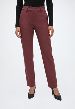 Femme portant un pantalon droit bordeaux taille haute, des chaussures noires à talons et un haut noir à manches longues, debout devant un fond uni.