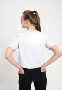 Žena s hnědými vlasy v culíku, oblečená v jednoduchém bílém tričku a černých džínách, se zády k objektivu, s rukama schovanými v zadních kapsách, na bílém pozadí.