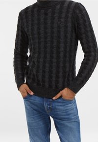 Pull noir en tricot avec motif torsadé, col montant, poignets et ourlet côtelés, porté avec un jean en denim bleu.