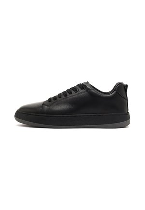 Zwarte lage leren sneaker met veters aan de voorkant, gewatteerde kraag en dikke zwarte rubberen zool op een witte achtergrond.