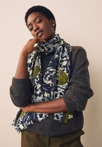 Écharpe à motifs bleu marine et crème avec des accents verts, drapée autour du cou, associée à un pull gris. Tissu doux et texturé.