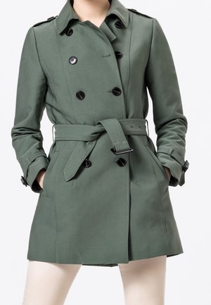 Femme portant un trench-coat vert à double boutonnage avec des boutons noirs et une ceinture nouée, les mains dans les poches, sur un fond uni.