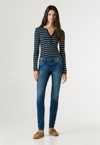 Noor naine kannab tumesinist triibulist pikkade varrukatega pluusi, siniseid kitsaid teksapükse ja beeže mokassiinjalanõusid, seisab ühevärvilise tausta ees.