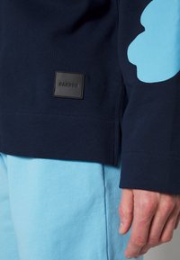 Felpa blu navy con una grande grafica azzurro chiaro sulla manica, presenta una toppa del marchio in pelle nera sull'orlo inferiore. Morbido tessuto di cotone.