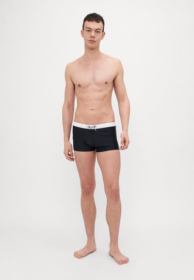 Shorts de bain noirs avec une ceinture à cordon blanche, au design ajusté et à la texture lisse, portés par une personne debout pieds nus.