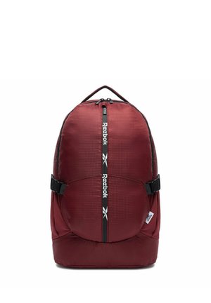 Vínově červený batoh Reebok se středním zipem zobrazujícím logo Reebok, černými bočními popruhy s přezkami a horním madlem pro nošení.