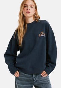 Granatowy sweatshirt z prążkowanym okrągłym dekoltem, luźny krój oraz haftowanym logo z wzorem słońca w odcieniach pomarańczowego, fioletowego i białego.