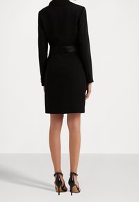 Černé blazerové šaty s dlouhými rukávy, přiléhavým střihem a saténovým páskem. Doplněné černými sandály na vysokém podpatku. Hladká textura.