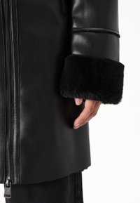 Musta nahkatakki, jossa on vetoketjukiinnitys ja tekoturkiskärjelliset hihansuut. Hihansuiden reunat ovat pehmeät, mikä kontrastoi takin sileää materiaalia.