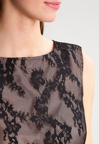 Svart spetsklänning med blommönster, genomskinlig textur och båtkrage. Närbild som fokuserar på halsringningen och detaljerna.