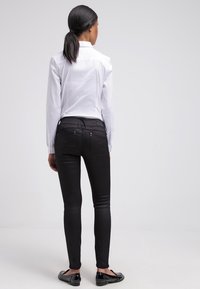 Jean ajustados negros vistos desde la parte de atrás, hechos de material elástico, con dos bolsillos traseros y detalles de costura sutiles. Combinados con una camisa blanca.